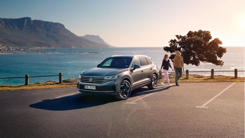 Ein VW Touareg Elegance parkt in der Sonne, Ansicht von schräg vorne, dahinter sind das Meer und die Küste zu sehen.
