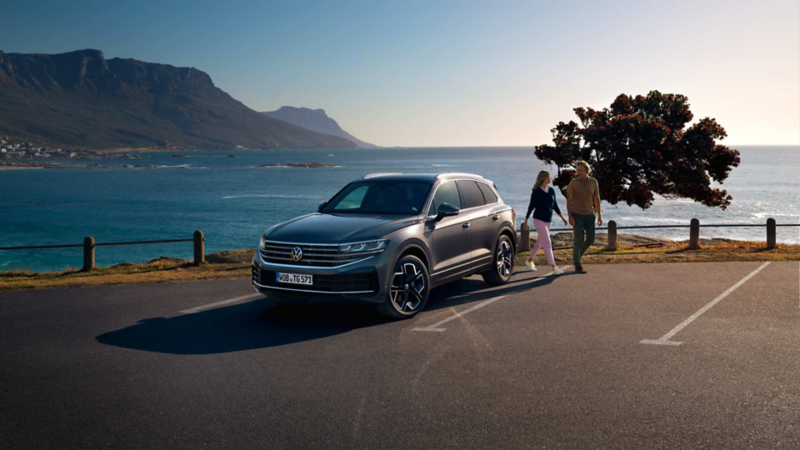 Ein VW Touareg Elegance parkt in der Sonne, Ansicht von schräg vorne, dahinter sind das Meer und die Küste zu sehen.