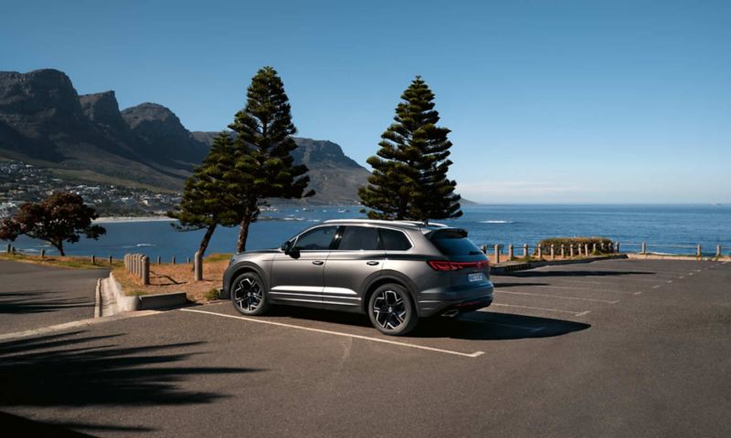 A VW Touareg Elegance parked in the sun, view from the side, behind it you can see the sea and the coast.