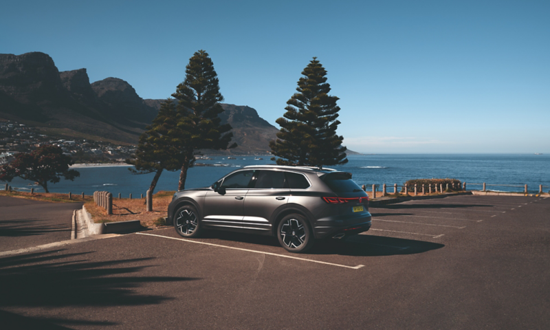 Een VW Touareg Elegance geparkeerd in de zon, vanaf de zijkant gezien, erachter zie je de zee en de kust.