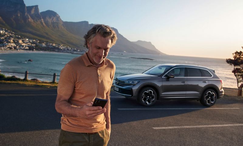Een man kijkt op zijn smartphone. Achter hem staat een VW Touareg Elegance geparkeerd, met de zee en de kust als achtergrond.