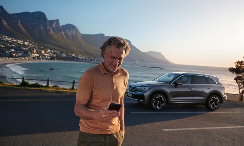 Mężczyzna patrzy na swój smartfon, za nim stoi zaparkowany VW Touareg Elegance, w tle morze i wybrzeże.