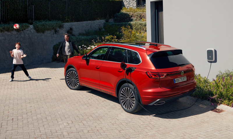 Un VW Touareg Elegance eHybrid, vue en diagonale arrière, est en charge devant une maison contemporaine.