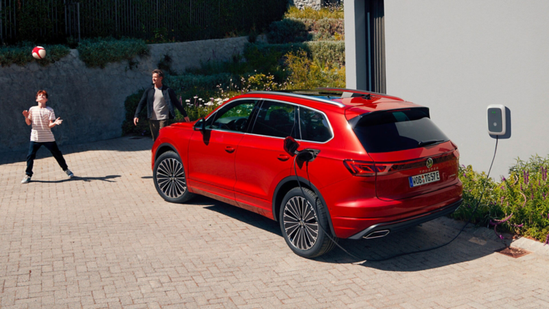 Un VW Touareg Elegance eHybrid, vue en diagonale arrière, est en charge devant une maison contemporaine.