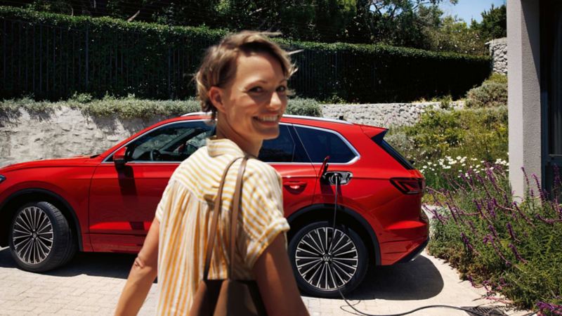 Una chica sonriendo delante de un Volkswagen Touareg rojo conectado a un cargador en casa