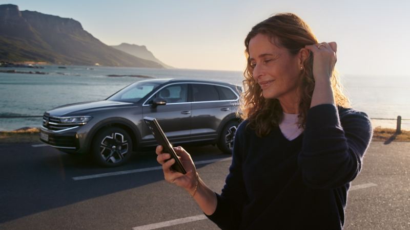 Vy av en leende person med en mobil i handen, i bakgrunden havet och en Touareg 