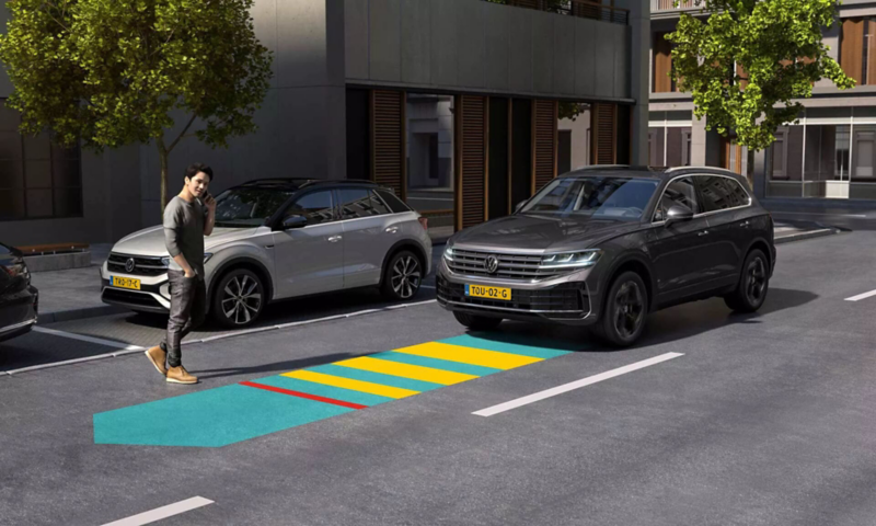 A man crosses a road, the Front Assist of the approaching VW Tiguan detects the pedestrian and warns the driver.