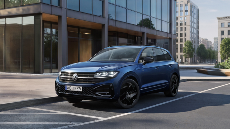 Ein blauer VW Touareg FINAL EDITION  steht vor einem Bürogebäude mit Glasfassade - Ansicht von schräg vorne.
