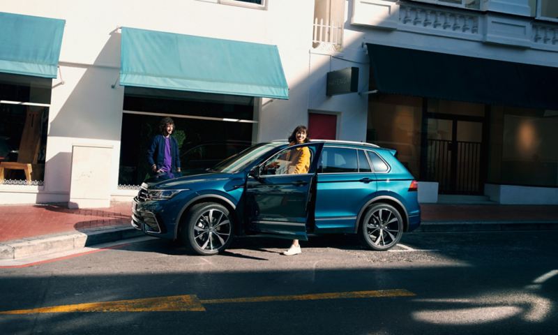 Blauer VW Tiguan in Seitenansicht steht am  Straßenrand, eine junge Frau steigt auf der Fahrerseite aus.