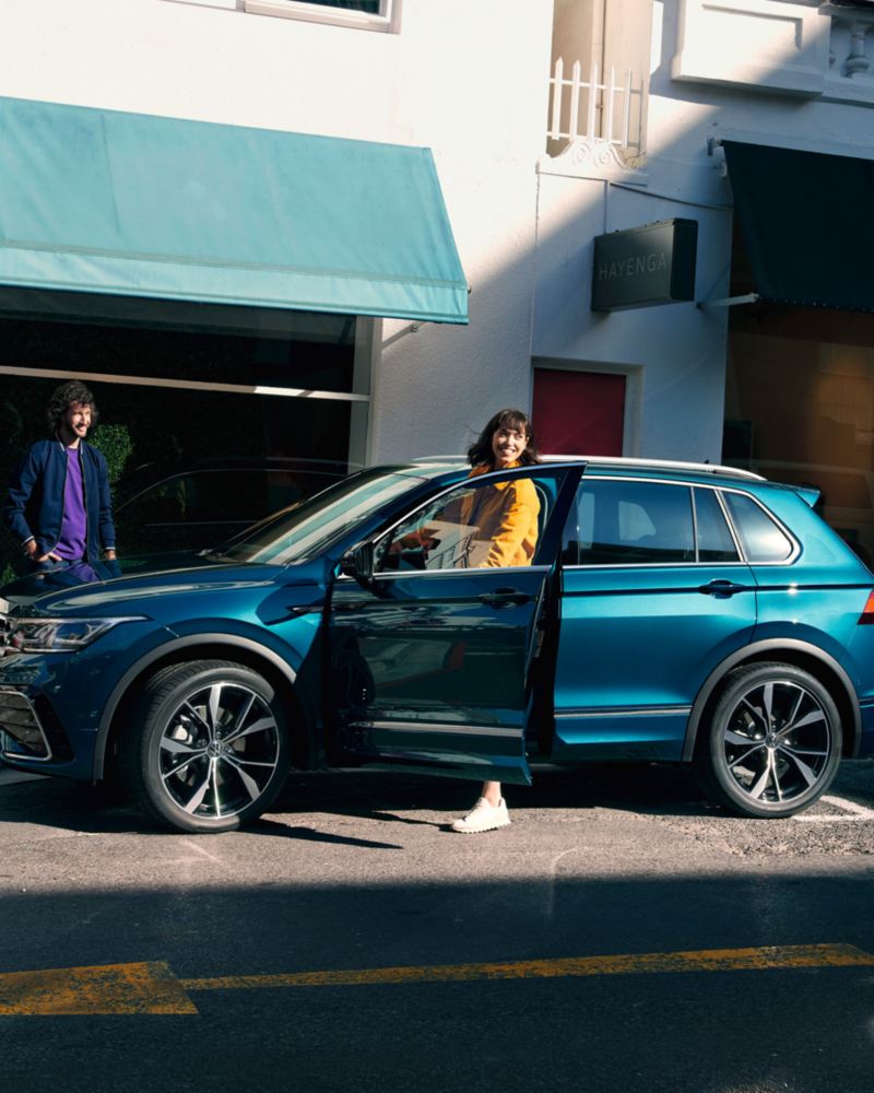 Blauer VW Tiguan in Seitenansicht steht am  Straßenrand, eine junge Frau steigt auf der Fahrerseite aus.