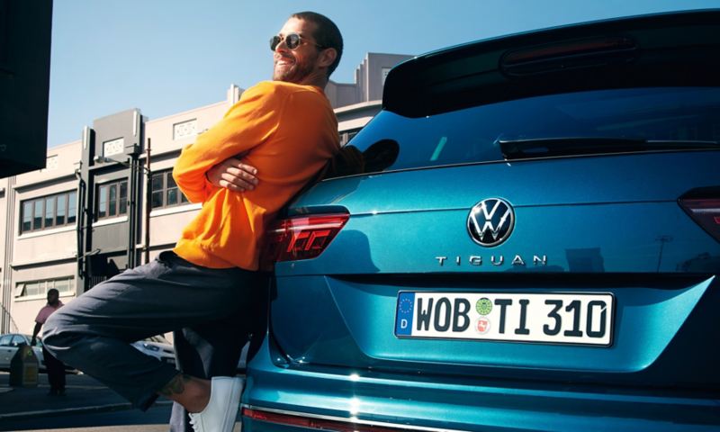 En glad mand iført solbriller læner sig op ad bagenden på en VW Tiguan.