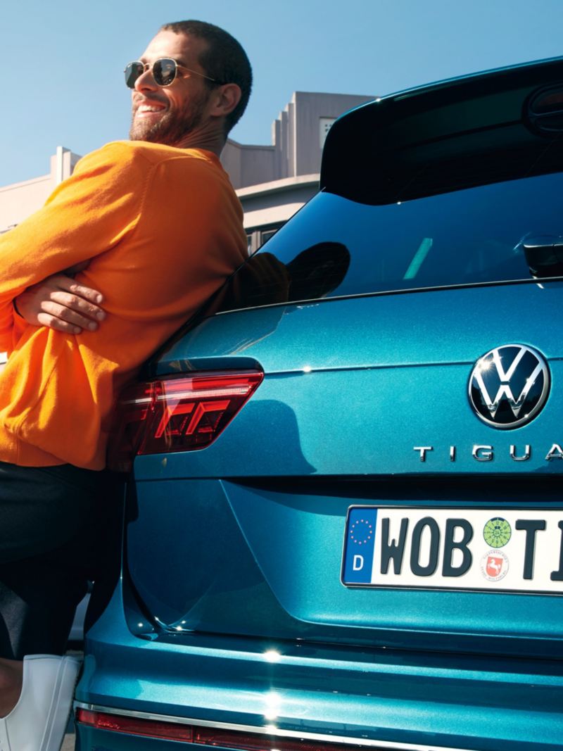 Mann mit Sonnenbrille lehnt glücklich am Heck eines blauen VW Tiguan.