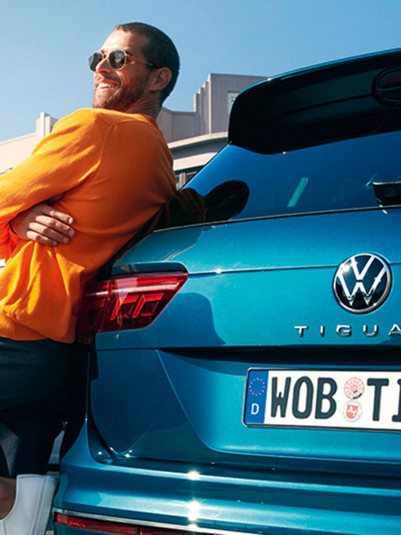 Un uomo con gli occhiali da sole si appoggia felicemente al retro di una VW Tiguan blu.