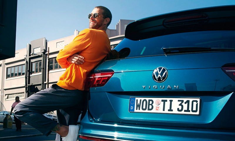 Man in sunglasses leans happily against the rear of a blue VW Tiguan.