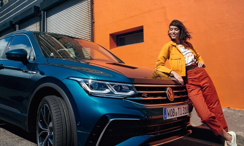 Donna appoggiata al cofano di una VW Tiguan blu, sorridente. Numerose funzionalità di bordo potranno essere attivate successivamente con Upgrades.