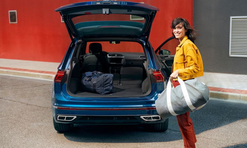 Una donna con una borsa sportiva sorride davanti al cofano del bagagliaio aperto di VW Tiguan blu.