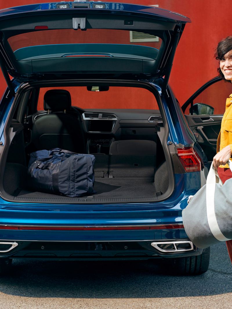 Woman with sports bag stands smiling at the open boot lid of the blue VW Tiguan.