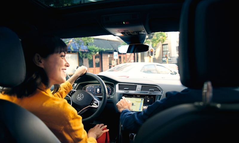 En kvinna vid ratten på en VW Tiguan med digital mätartavla, passageraren sträcker ut sin hand mot navigationssystemet.