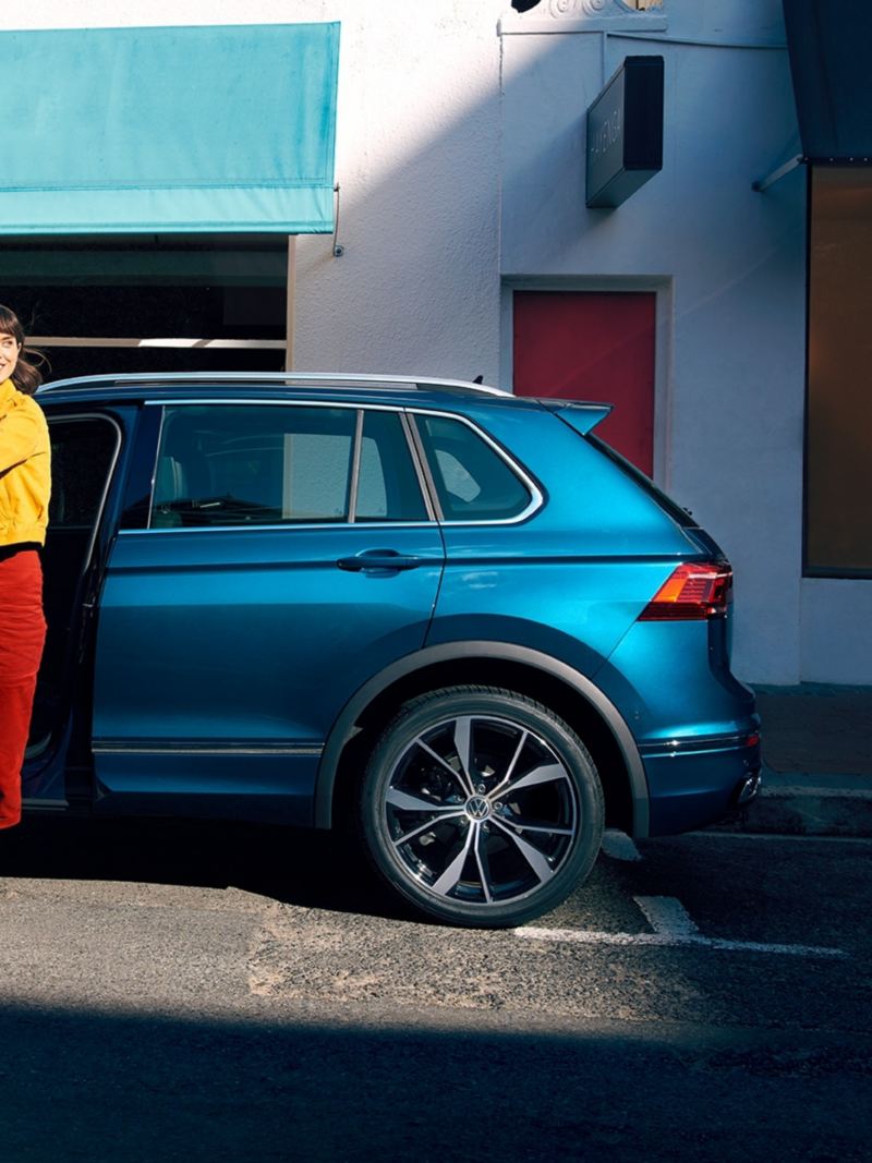 Blauer VW Tiguan in Seitenansicht steht am Straßenrand, eine junge Frau steigt schwungvoll auf Fahrerseite aus.