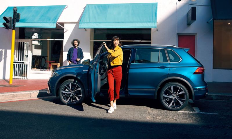 Blue VW Tiguan viewed from the side, standing at the roadside, a young woman gets out of the car energetically on the driver’s side.