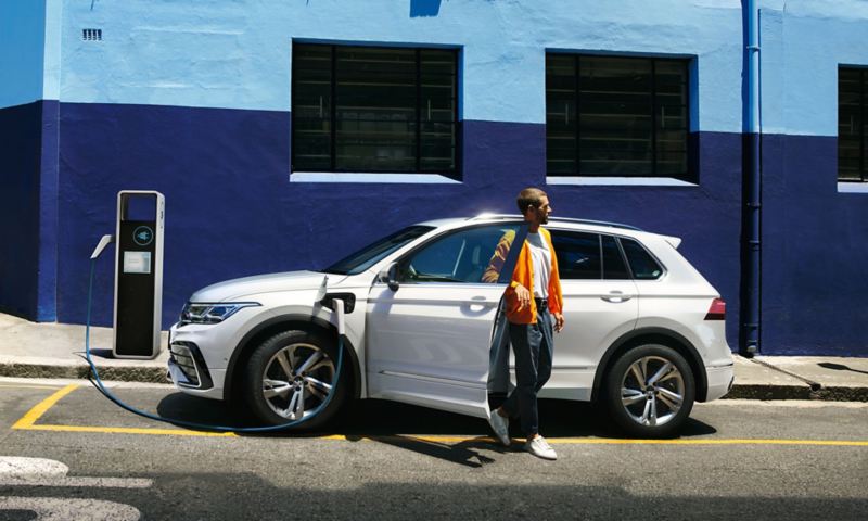 VW Tiguan eHybrid in white stands in front of a blue building on the roadside and charges at a charging station, a man gets out.