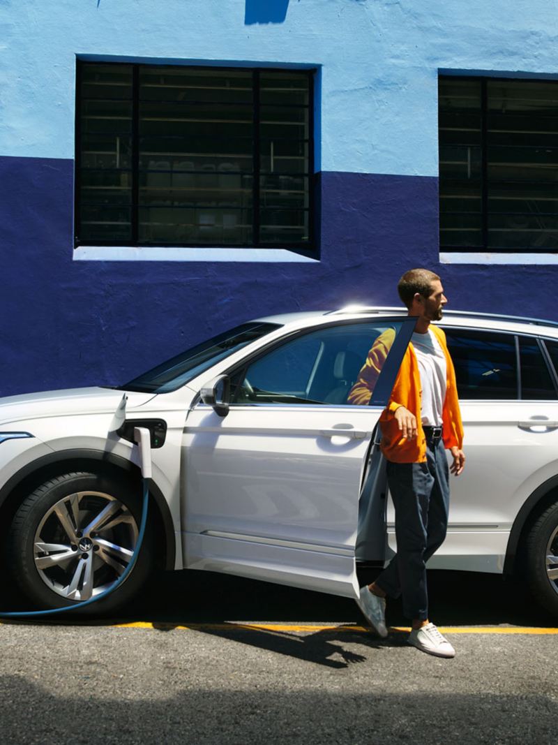 The new Tiguan eHybrid  Side view of the VW Tiguan eHybrid in white, which charges at the roadside. Young man gets out of the driver's side.