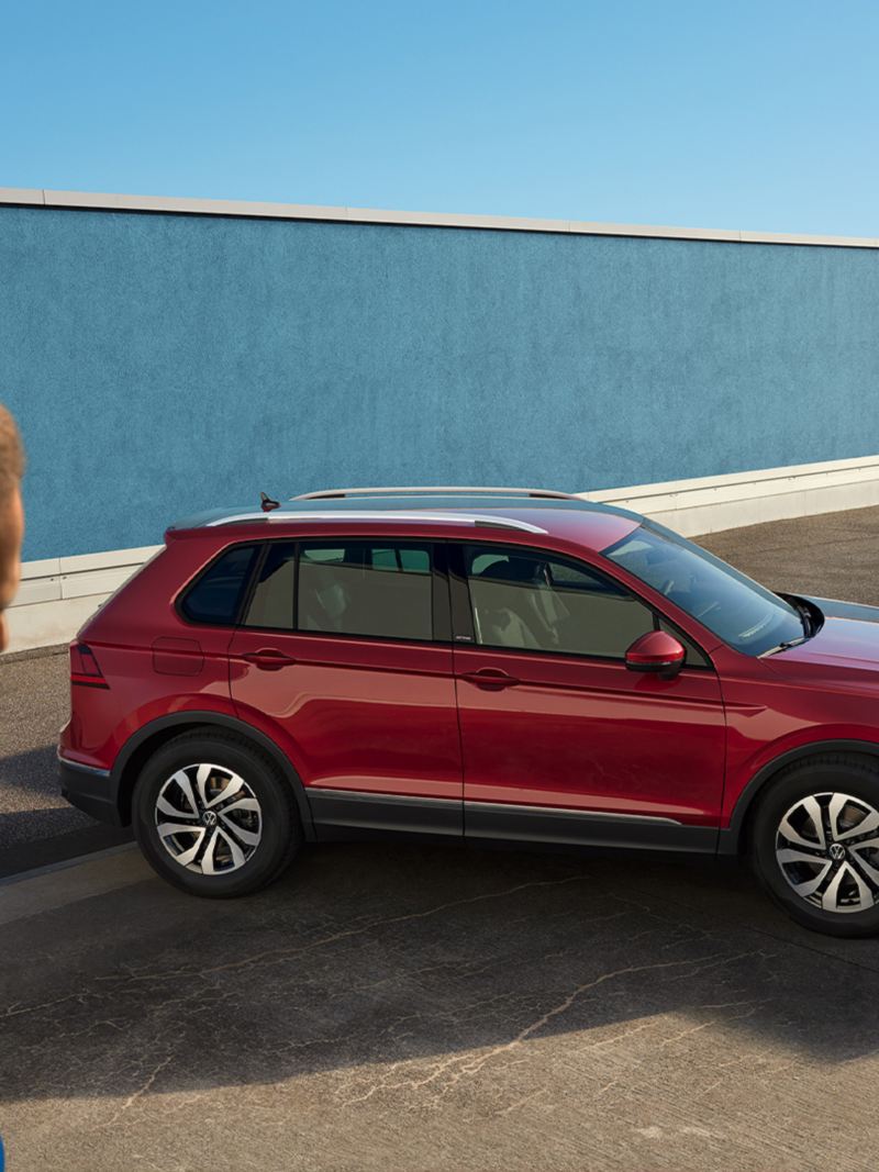 Le VW Tiguan rouge se tient dans un environnement urbain devant un mur bleu. Il y a un homme sur la gauche de la photo.