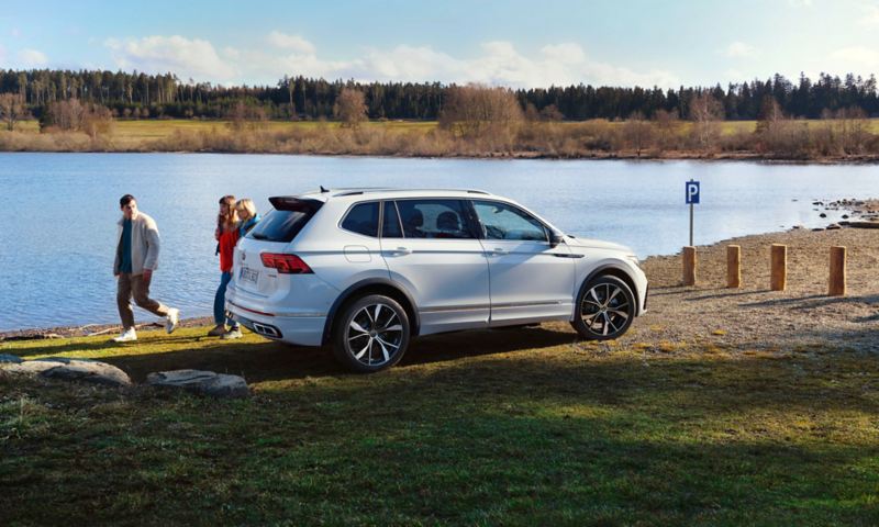 A group of young people walk past a white Tiguan Allspace R-Line parked by a lake.