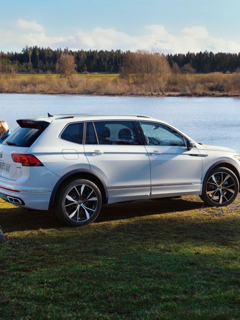A group of young people walk past a white Tiguan Allspace R-Line parked by a lake.