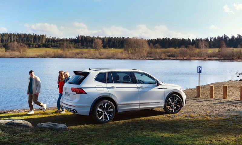 Een groep jongeren loopt langs een witte Tiguan Allspace R-Line die bij een meer geparkeerd staat.