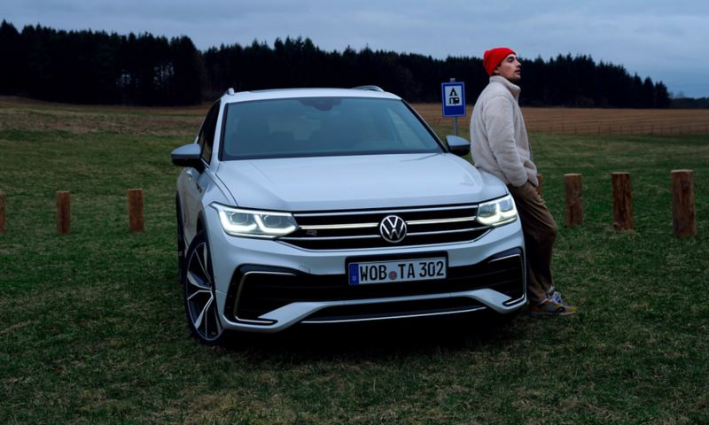 A man leans against the hood of a white Tiguan Allspace R-Line, the LED matrix headlights and light bar on. 