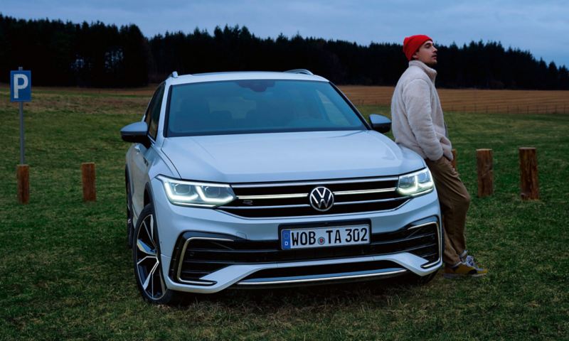 Un uomo è appoggiato al cofano di una Tiguan Allspace R-Line bianca, i fari LED Matrix opzionali e il listello luminoso sono accesi. 