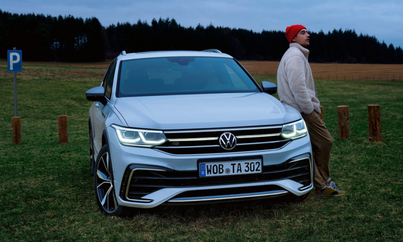 Ein Mann lehnt an der Motorhaube eines weißen Tiguan Allspace R-Line, die optionalen LED-Matrix-Scheinwerfer und Lichtleiste sind an. 