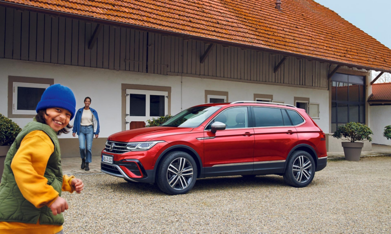 Vista laterale di una Tiguan Allspace rossa in un maneggio. Una donna in piedi davanti all’auto, un bambino corre ridendo.