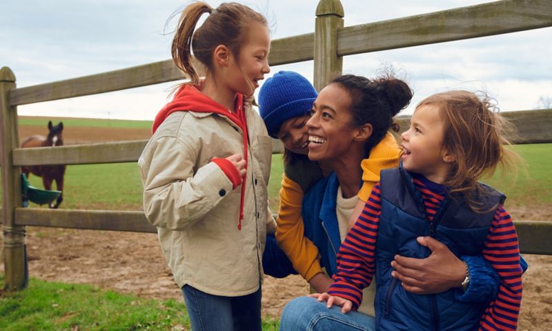 Eine Frau mit drei Kindern hockt vor dem Zaun einer Pferdekoppel, sie lachen. 