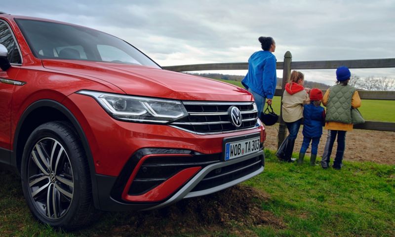 Side view of the front of a red Tiguan Allspace; in the background a woman is standing with three children at the fence of a paddock. 
