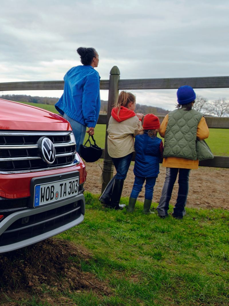 Side view of the front of a red Tiguan Allspace, in the background a woman with three children is standing at the fence of a horse pasture. 