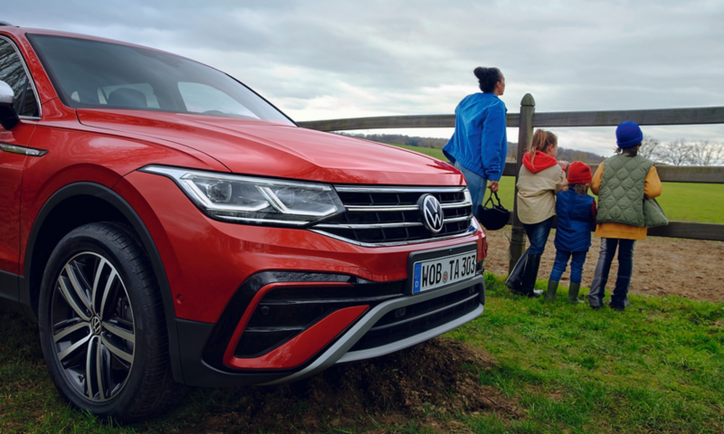 Side view of the front of a red Tiguan Allspace, in the background a woman with three children is standing at the fence of a horse pasture. 