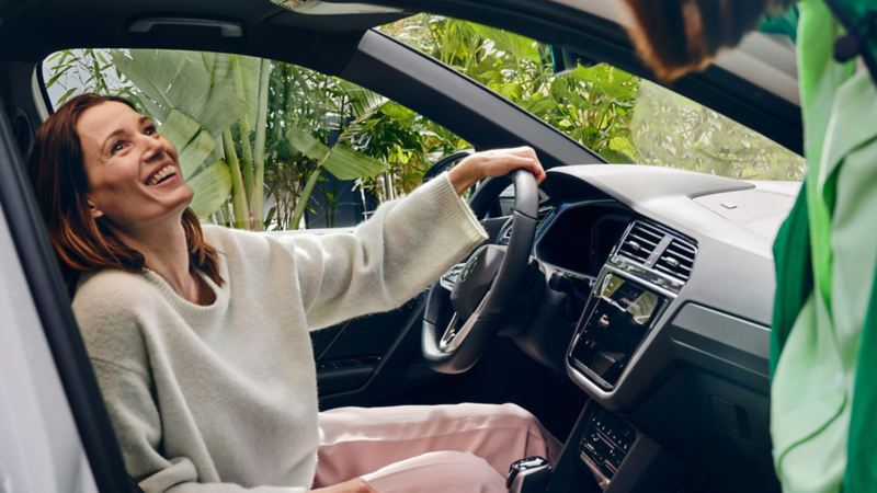Una donna siede sorridente sul sedile del guidatore di una Volkswagen Tiguan eHYBRID, tenendo la mano sinistra sul volante.