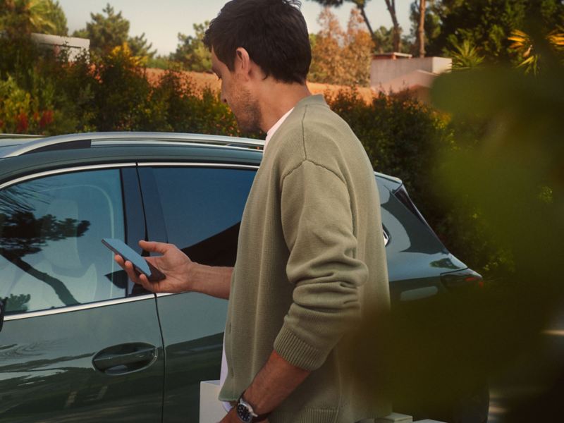 Asmuo stovi priešais „Volkswagen“ automobilį ir laiko rankose mobilųjį telefoną