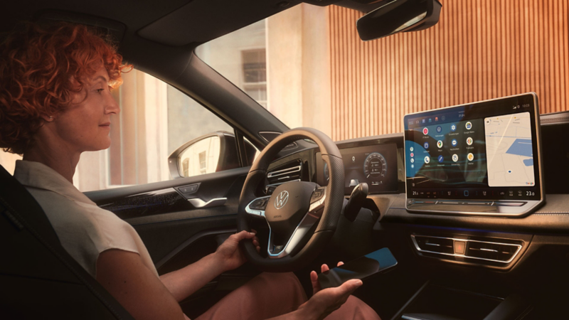 Μία γυναίκα οδηγός μέσα στο VW Tayron. Κρατάει το κινητό τηλέφωνο το οποίο είναι συνδεδεμένο με το App-Connect στο αυτοκίνητο και βλέπουμε την οθόνη Infotainment με τις εφαρμογές.
