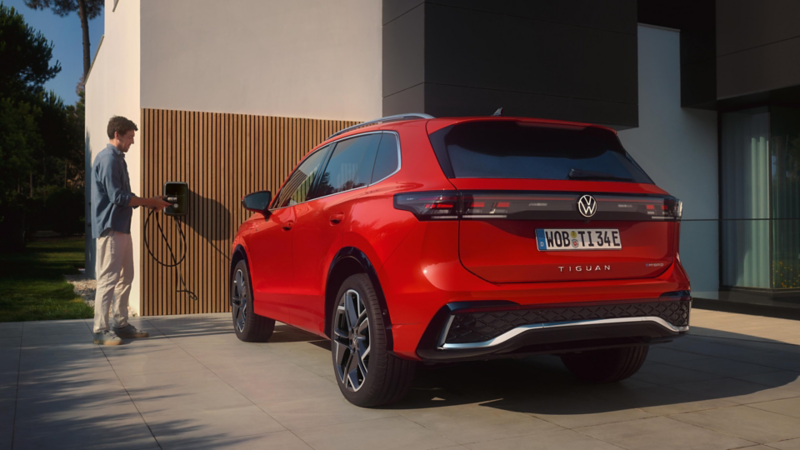 Ein roter VW Tiguan eHybrid, der vor einem Haus parkt, wird von einem Mann über die Wallbox am Haus geladen.