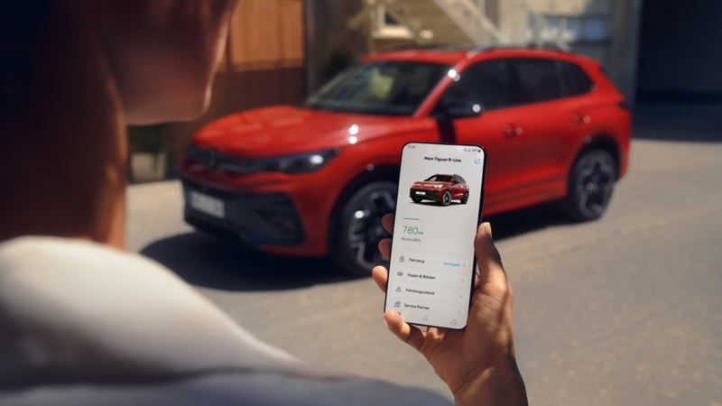 Blick auf ein Handydisplay mit Fahrzeugdaten des Tiguan, im Hintergrund ein seitlich geparkter VW Tiguan in rot