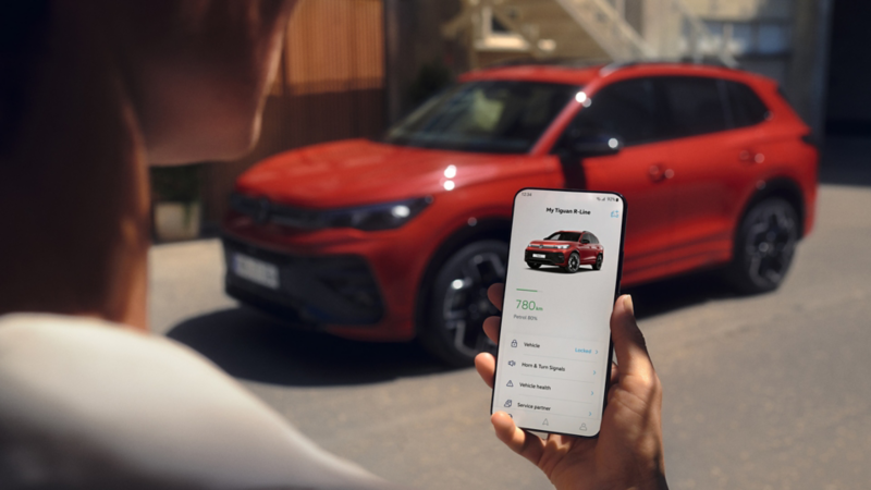 Skats pār kāda cilvēka plecu uz mobilo tālruni, kurā ir atvērta Volkswagen lietotne. Fonā redzams sarkans Tiguan R-Line.