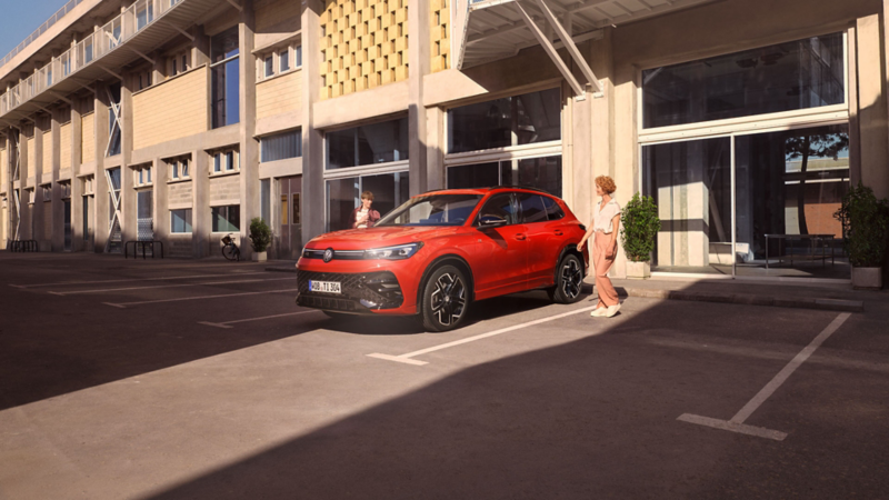 Roter VW Tiguan steht vor einem Gebäude mit Balkonen, eine junge Frau geht auf die Fahrerseite zu.