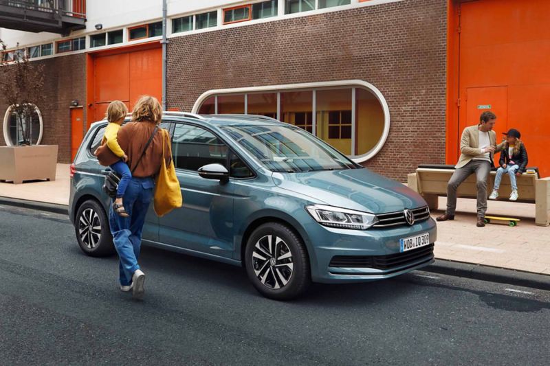 Una donna con in braccio un bambino si avvicina alla sua Volkswagen Touran, vista lateralmente parcheggiata a bordo strada.