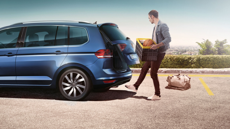 Mann mit Kiste öffnet per „Easy Open und Close“ die Heckklappe eines blauen VW Touran.