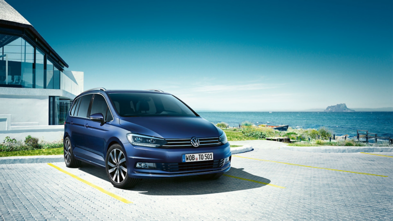 En blå VW Touran holder foran en indkørsel, en familie med to børn og en hund kommer ud fra det moderne hus.