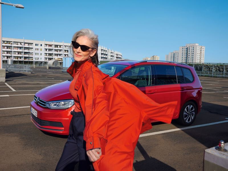 Roter VW Touran ACTIVE auf urbanem Parkplatz. Stadt im Hintergrund. Blick auf Leuchten und Seite. Davor Frau mit roter Jacke.