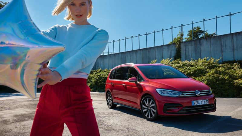 VW Touran rouge ACTIF dans un parking urbain. Ville en arrière-plan. Vue des lumières et de l'avant. Une femme en veste rouge devant.